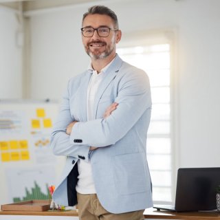 Man with glasses and a suit stands in his office and smiles 