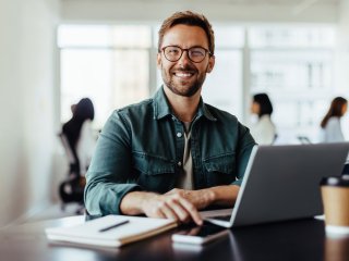Mann mit Brille und braunen Haaren sitzt an seinem Schreibtisch und hat seinen Laptop, sein Handy und einen Block vor sich