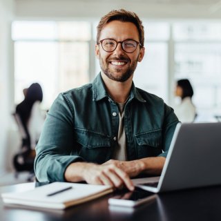 Man with glasses and brown hair sits at his desk with his laptop, cell phone and a notepad in front of him