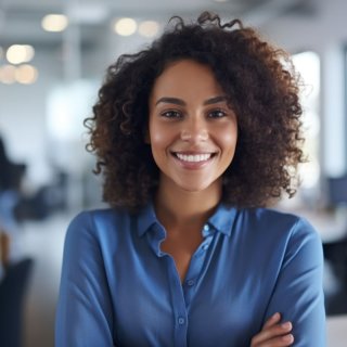 Woman with long brown curls stands in an open-plan office and smiles