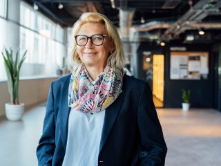 Frau mit langen blonden Haaren steht in einem Unternehmensflur und lächelt